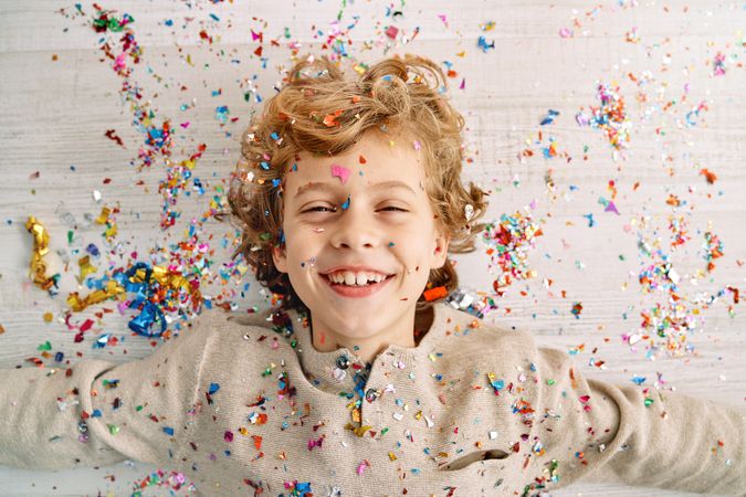 Joyful boy lying on wooden floor covered with confetti