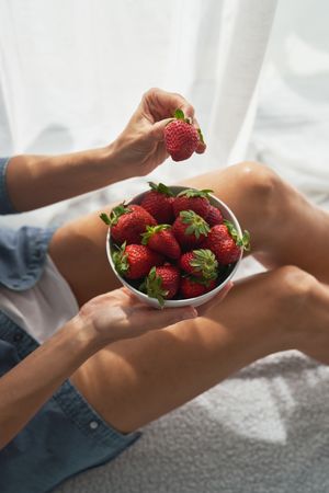 Unrecognizable female taking strawberry from bowl while sitting