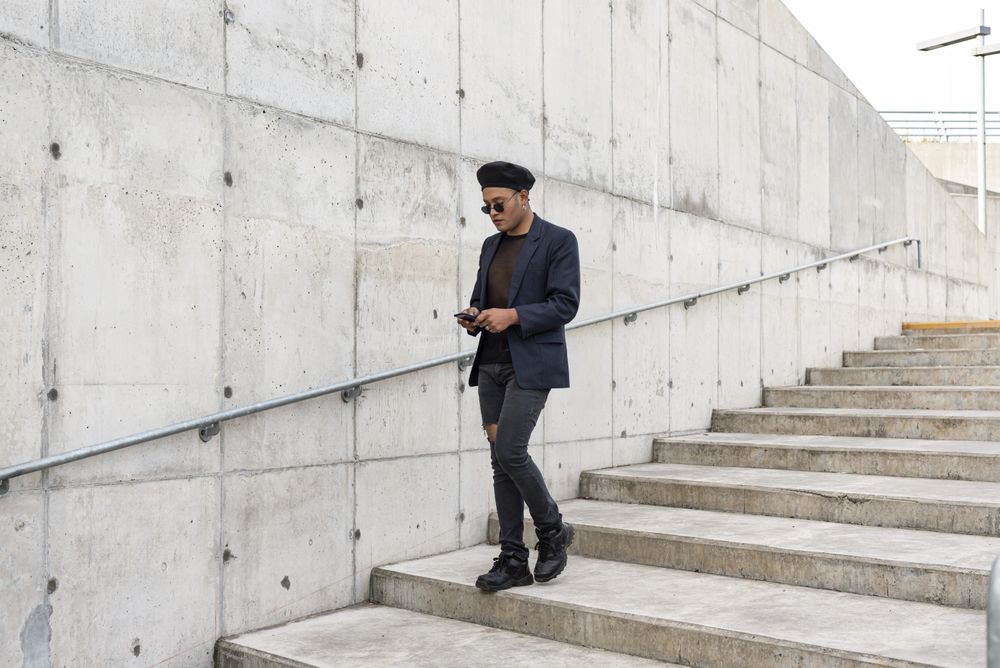 Young Latino gay man wearing a fashionable hat and sunglasses, walking down the stairway while texting on his smartphone