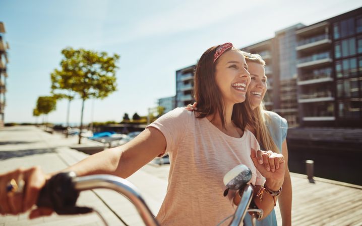 Two laughing female friends with arms on each others shoulders while walking with bike next to river