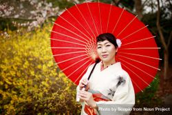Portrait of Japanese woman in light kimono holding a red umbrella standing near yellow meadow 5XARG4