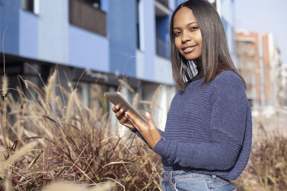 Confident female in sweater standing outside blue building in long grass with tablet
