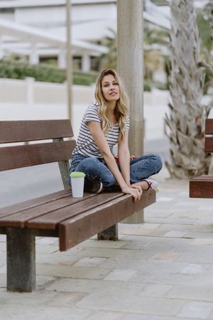 Confident woman with disposable cup of coffee sitting on street bench