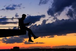 Out line of male sitting on ledge with tablet against a colorful dusk side, vertical