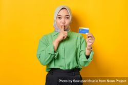 Smiling Muslim woman in headscarf and green blouse saying “shhh” and holding her credit card 4NLp90