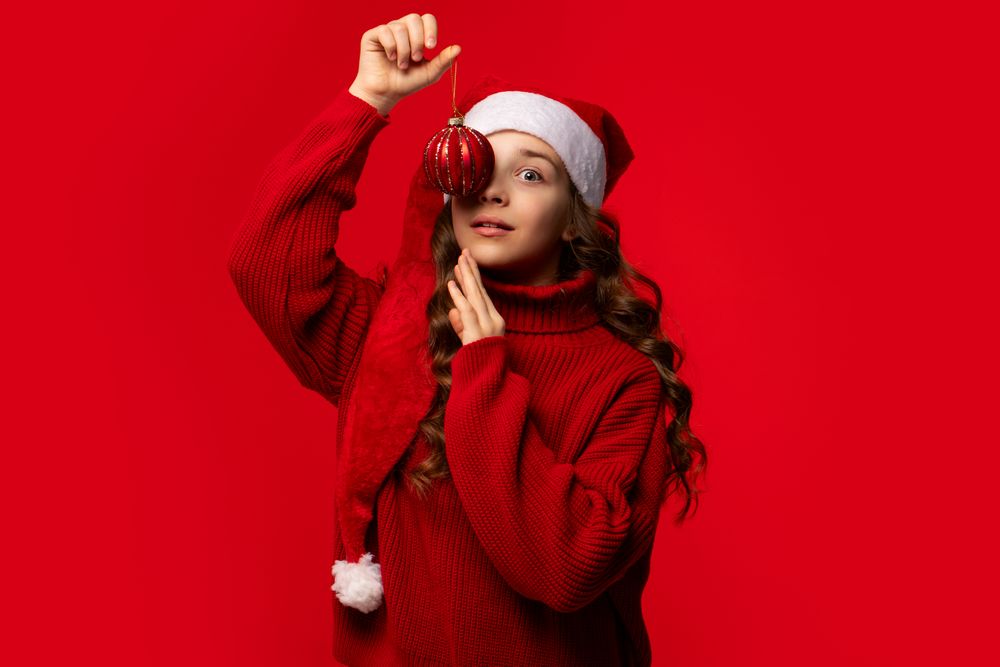 Girl in Santa hat holding red Christmas ornament near eye.