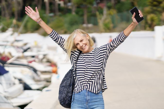 Older woman in jeans and striped shirt standing outside on sunny day with arms up