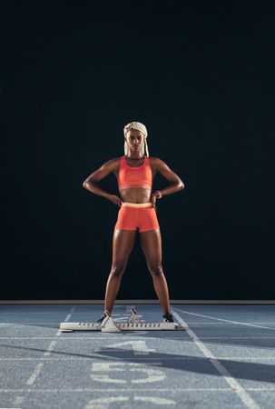 Female athlete standing beside a starting block on running track on a dark background