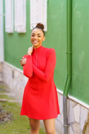 Smiling female in red zipped up dress standing outside green building