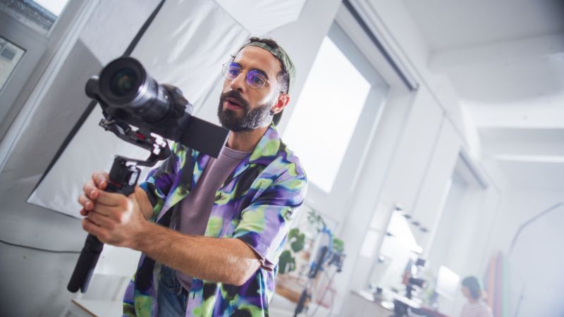 Focused Young Male Videographer With Beard, Filming In A Bright Studio, Holding A Professional Camera, Surrounded By Modern Filmmaking Equipment, Engaged In A Creative Production Process.