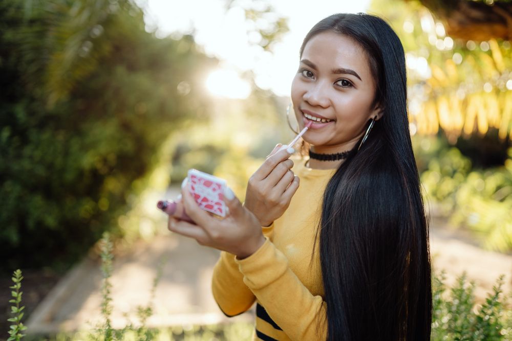 Stylish long haired Asian woman smiling and applying lip gloss