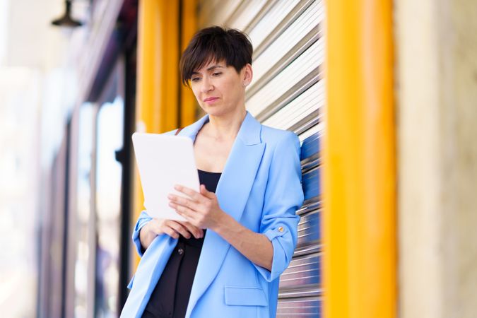 Woman in blue blazer leaning on shutter outside reading digital tablet