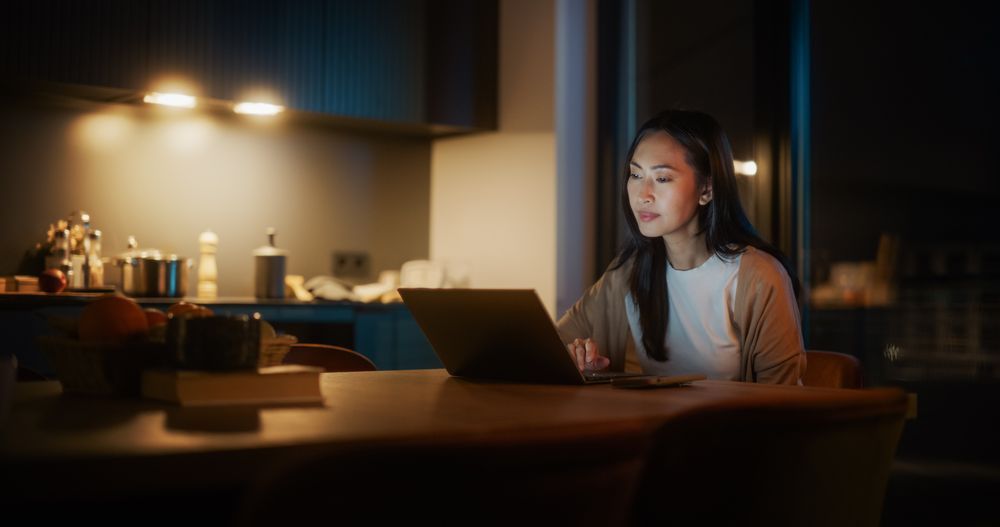 Authentic Asian Woman Sitting at a Table in Cozy Kitchen and Using Laptop Computer at Home at Night.