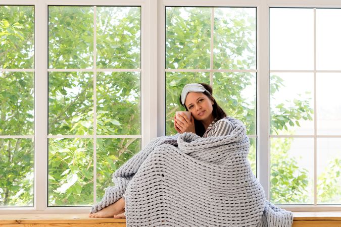 Relaxed woman sitting by window with mug and blanket.