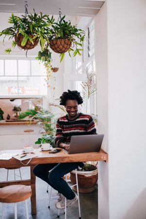Man smiling while working on his computer at a coffee shop with hangplants