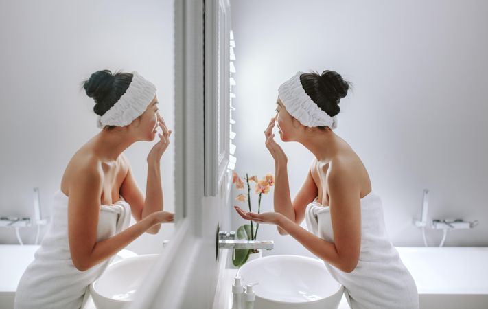 Woman wrapped in towel applying cream on face standing in front of mirror