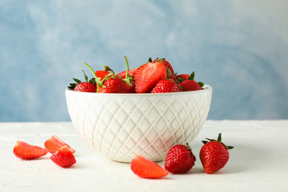 Bowl with fresh strawberries on table against blue background, space for text. Summer sweet fruits and berries