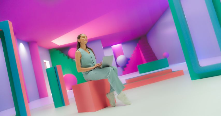 Young Woman in Bright Outfit Using Laptop Computer While Sitting in Geometric Abstract Environment. Creative Young Female Joyfully Checking Social Media in a Studio with a Colorful Background