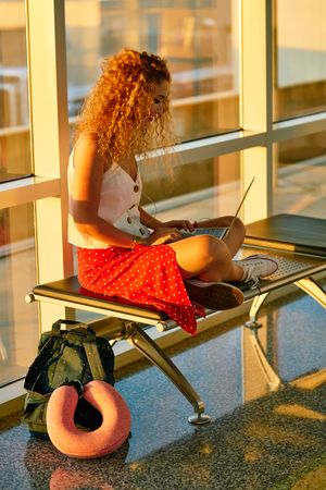 Enthusiastic woman working on laptop sitting on bench in airport hall
