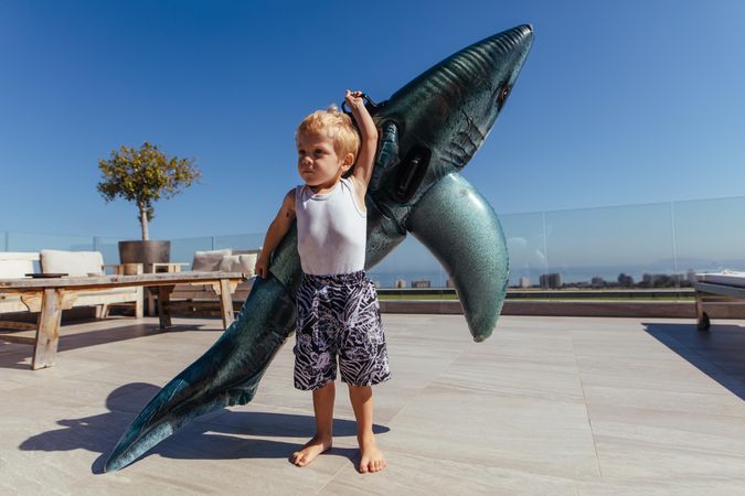 Little kid lifting a big inflatable toy shark by the poolside
