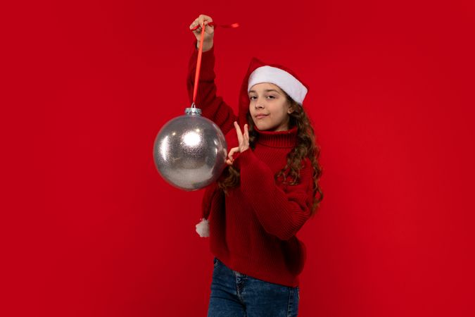 Playful girl in Santa hat and sweater holds Christmas ornament