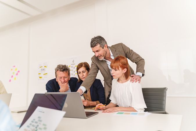 A group of colleagues engaged in a focused discussion around a laptop in a modern office, with work documents and sticky notes