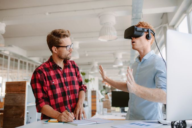 Businessman wearing VR goggles and colleague writing notes in office