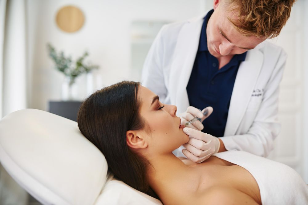 Smiling male dermatologist injecting treatment into brunette patient’s lips
