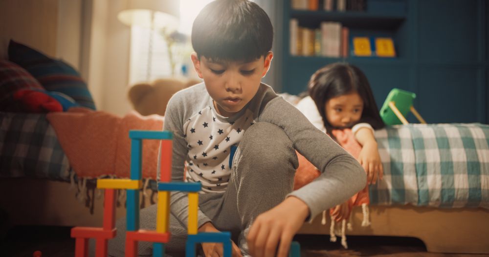 Portrait of Focused Smart Little Boy Playing with Construction Building Blocks in Children's Room, His Little Sister is Resting on a Bed.