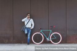 Male in sunglasses standing with red and green bicycle leaning on wall and using phone 0KAGY4