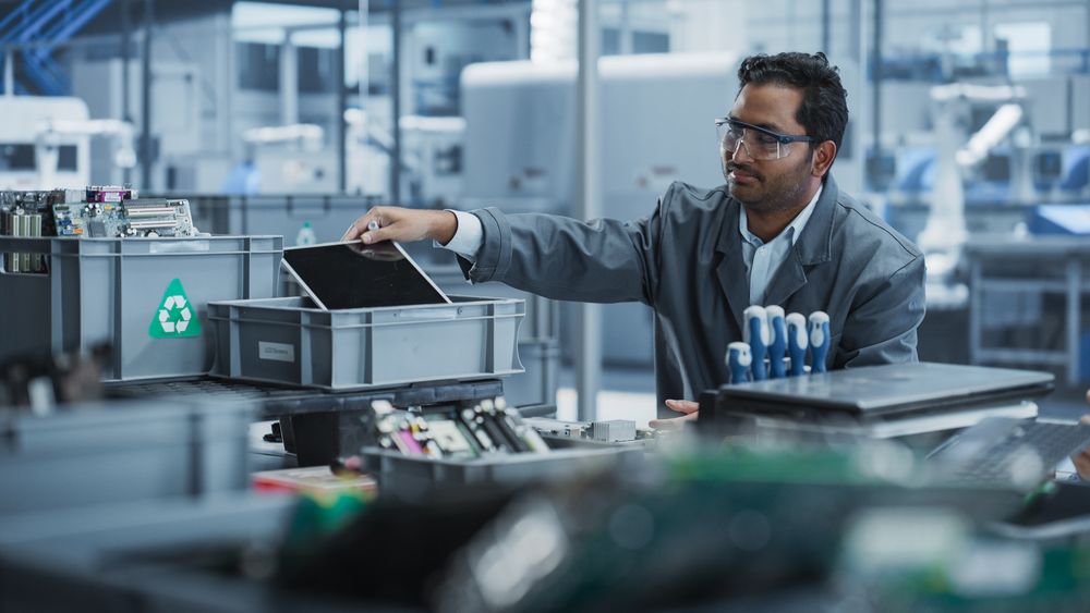 Indian Male Worker Disassembling Laptops To Recycle Electronic Components For Microchip Production At An Electronics Factory With Robotic Arms.