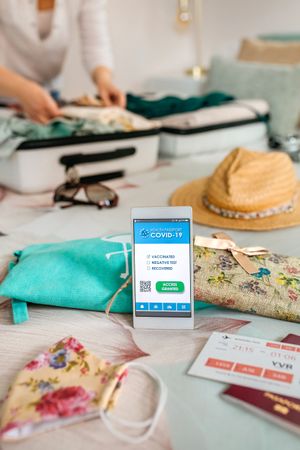 Mobile phone with covid passport with unrecognizable woman preparing suitcase for vacation