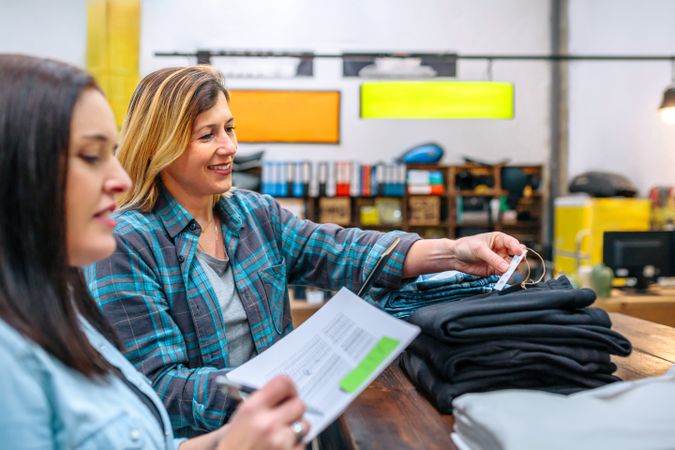 Happy woman checking price label of pants next to her colleague on second hand clothing store
