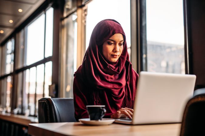 Muslim woman sitting at cafe table and working on laptop with coffee in side