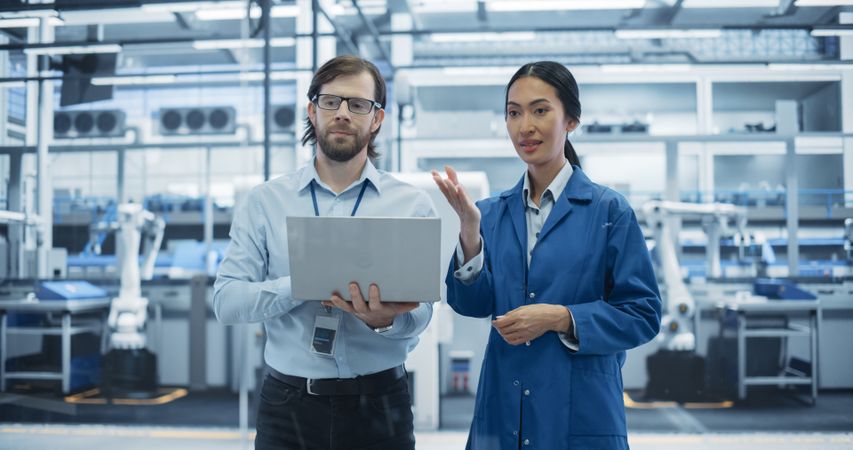 Man and Asian Female Having a Conversation at Industrial Facility. Advanced Factory Employees Working Together, Using Laptop Computer for Supervising Work Process in a Modern Facility