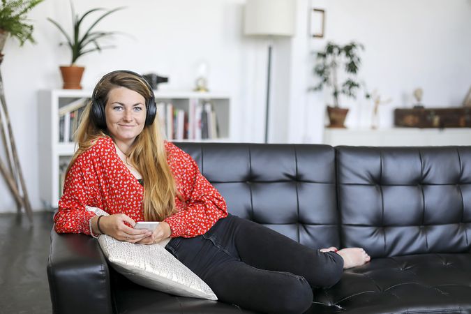 Happy woman listening to music on headphones looking at camera sitting on a sofa in the living room