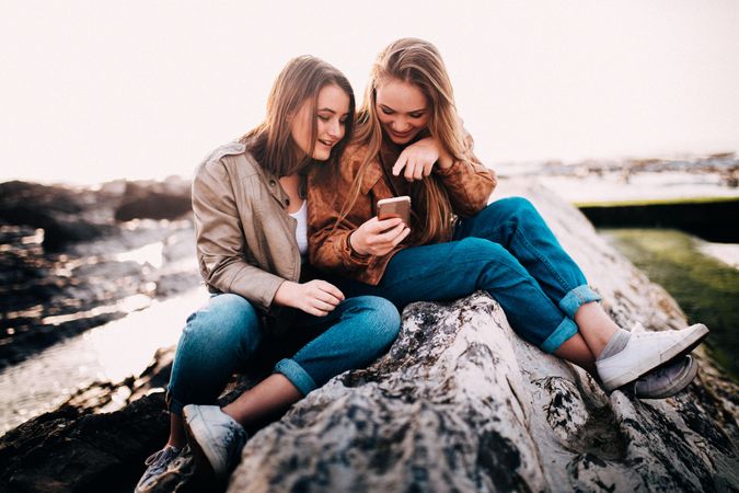 Two young women sitting on a rock outside looking at a phone