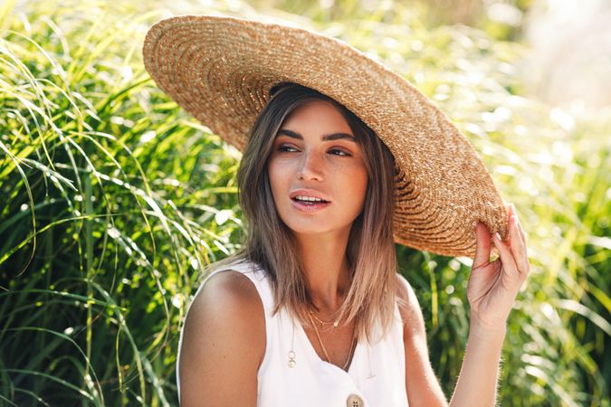 Woman sitting in front of long grass looking away with hand on her straw hat