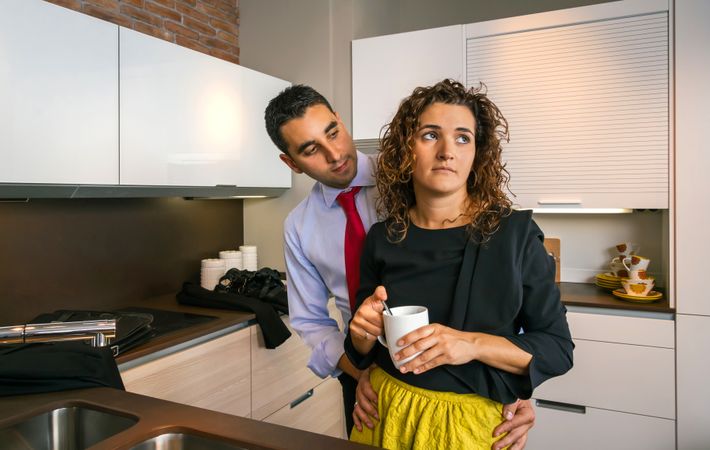 Businessman embracing annoyed curly woman while holding cup of coffee in the kitchen