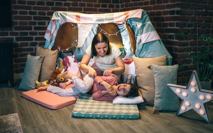 Woman tickling to little girl while laughing and shouting lying over cozy mats in night pajama party