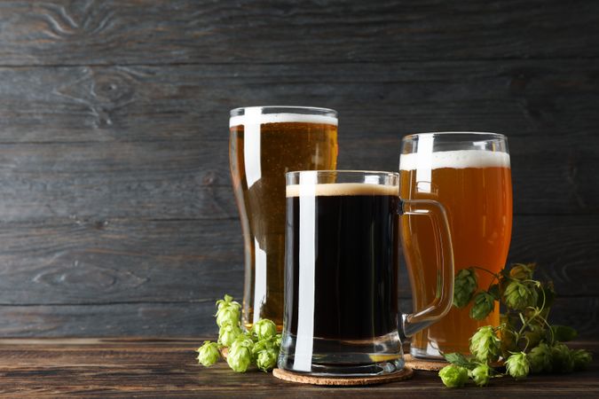 Different mugs with beer and hop cones on wooden table, copy space