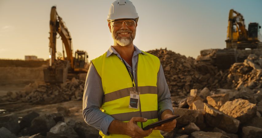 Successful Bearded Civil Engineer Wearing Protective Goggles And Smiling At Camera On a Construction Site on Sunny Day While Heavy Machinery Working. Man Holding Tablet, Wearing Hard Hat, Safety Vest.