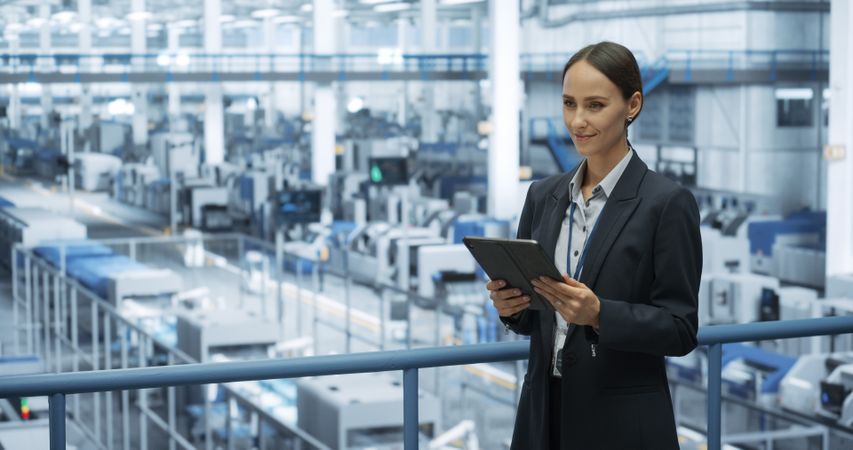 Portrait of a Happy Engineer Using Laptop Computer and Looking Around a Factory Facility with Automated Robotic Arms Producing Modern Electronic Components for Different Industries
