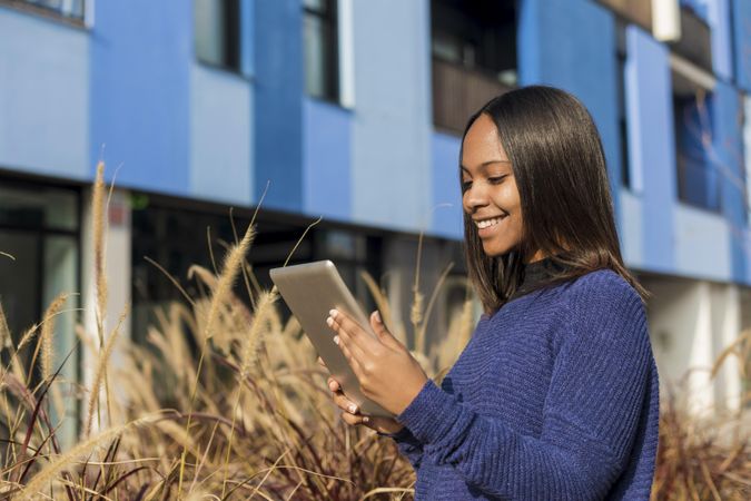 Happy female in sweater standing outside blue building with tablet