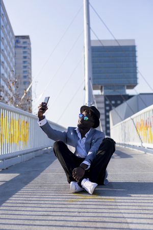 Black man in suit sitting on footbridge taking a selfie