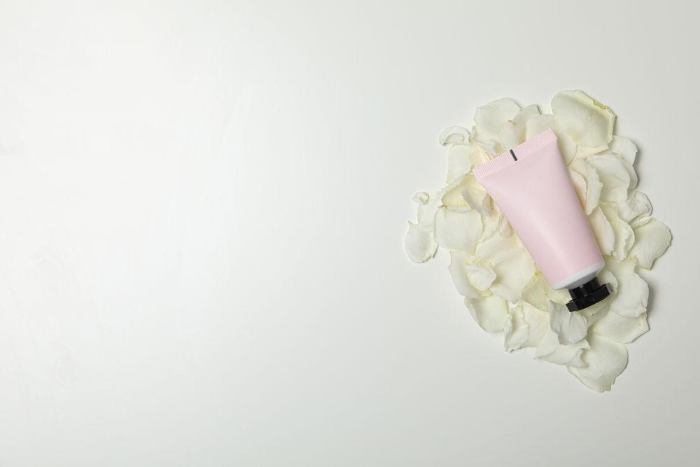 Tube with cream on rose petals on light background, space for text