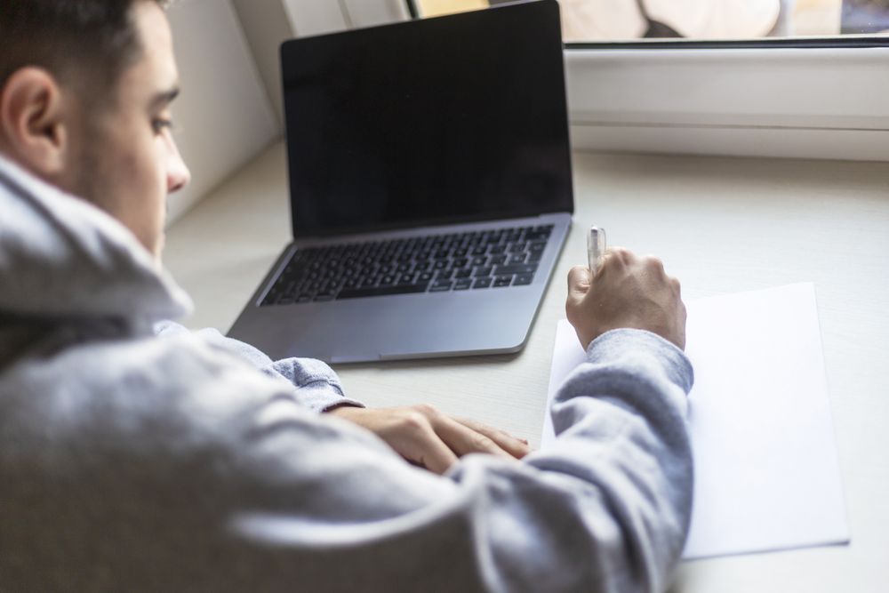 Rear view of a young man writing notes next to laptop at home