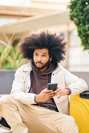 Young man with curly afro hair in the street sitting with the phone in his hand looking around him