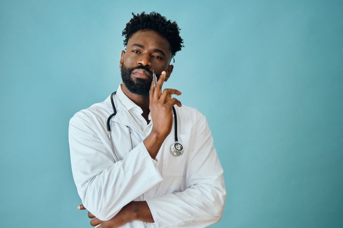 Thoughtful Black male doctor in blue studio with pen to his chin