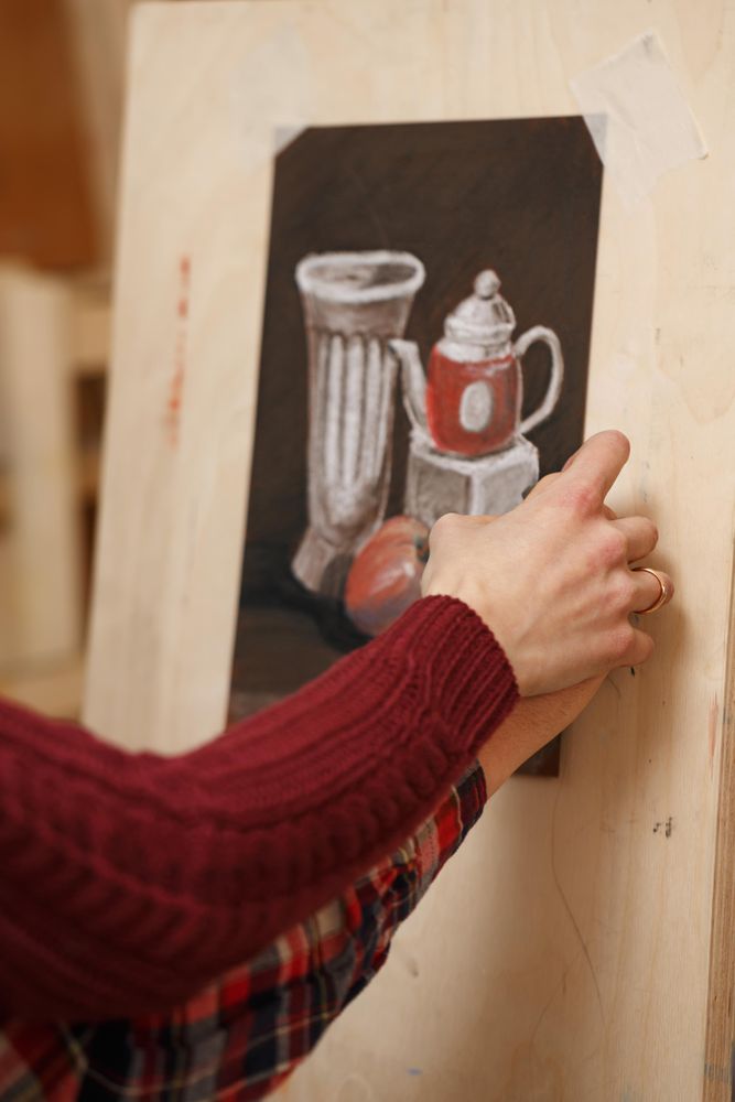 Hand of teacher guiding student as he draws a still life of tea pot